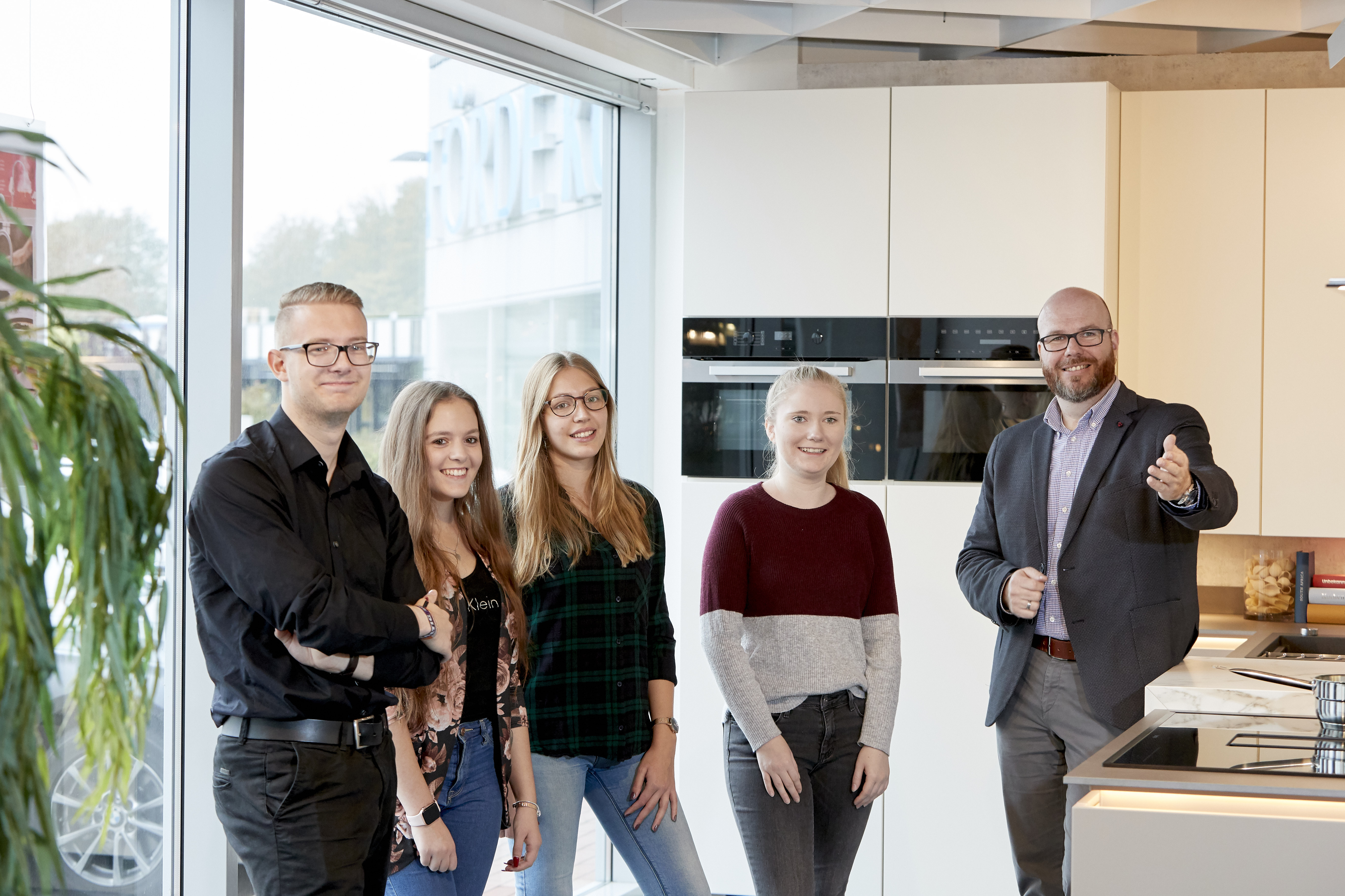 Auf diesem Bild sieht man 4 Auszubildende und einen Ausbilder in der Flensburger Förde-Küchen Filiale. Sie stehen gemeinsam in einer Einbauküche und freuen sich über den Fototermin.
